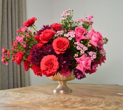 Flower arrangement composed of various shades of fuchsia using roses, cushion pompons, sparkz and inconfetti. arrangement is placed inside a silver vase on top a wooden table
