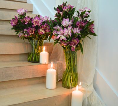 Two arrangements of purple Astronova in tall clear vase sitting on wooden staircase steps showcasing the flowers long green stems. Near the vases are three white lit candles, one on each step.
