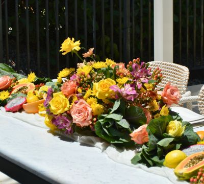 Outdoor flower arrangement with yellow, purple, peach, and pink roses. Local tropical fruits like lemons, papayas, dragonfruit, grapefruit, and oranges are displayed across table surrounding flower arrangement.
