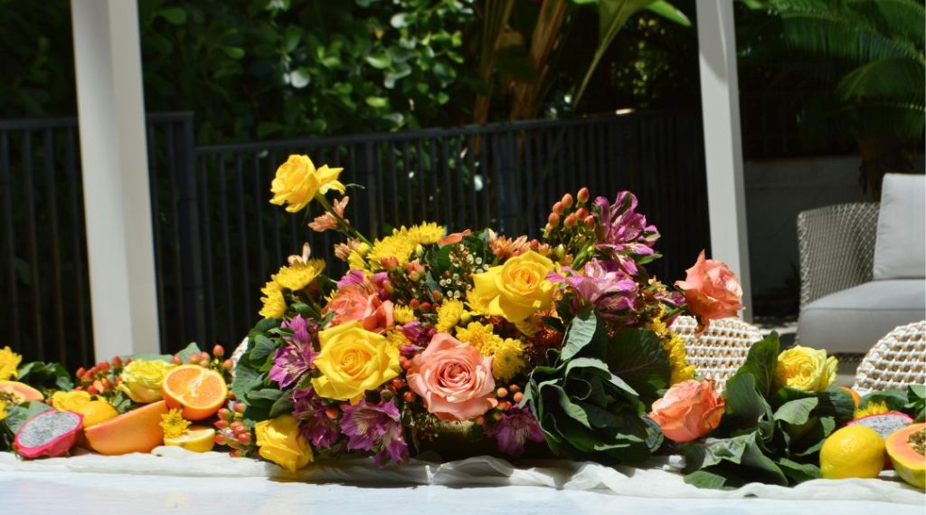 Outdoor flower arrangement with yellow, purple, peach, and pink roses resembling a fruit and vegetable wedding trend.