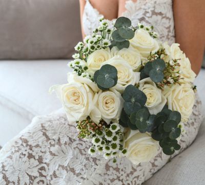 Women in white lace wedding dress hold round bouquet of white roses with touches of eucalyptus and inconfetti whilst sitting down.