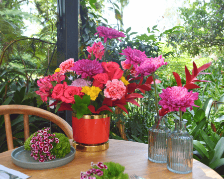 Lush red and pink flower arrangement in gold-trimmed red vase with garden backdrop