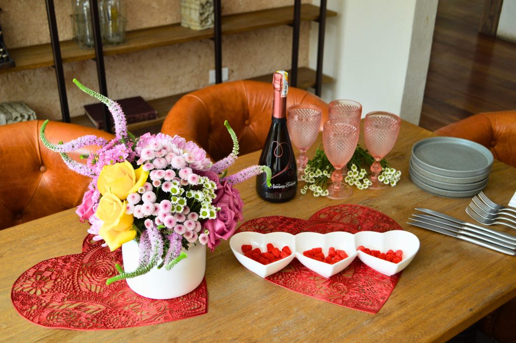 Valentine's Day table setting with pink and yellow floral arrangement, red heart placemats, and pink champagne glasses