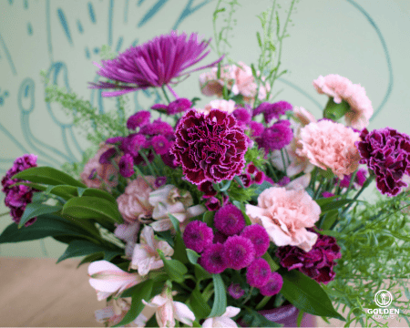 Close-up of vibrant floral arrangement featuring fuchsia spider mum, burgundy button flowers, pink carnations and white alstroemeria against light blue background