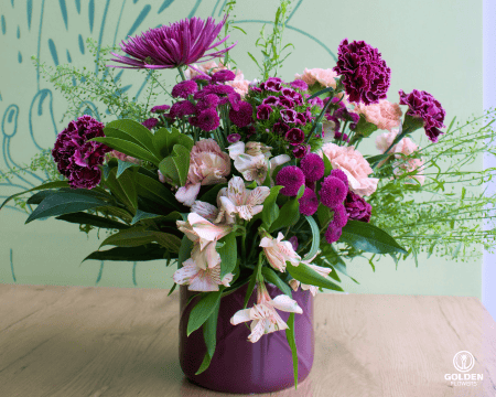 Elegant floral mothers day arrangement with burgundy chrysanthemums, pink carnations and white alstroemeria in a purple vase against a mint green wall with botanical line art