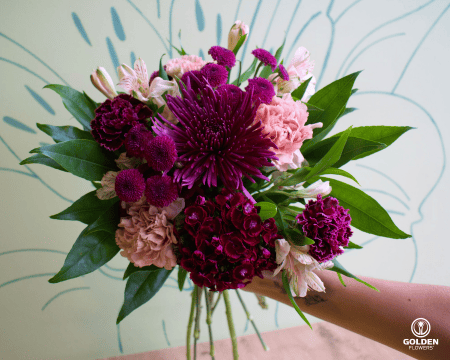Hand holding a hand-tied bouquet of magenta chrysanthemums, burgundy carnations, pink carnations and white alstroemeria against mint green wall with floral outline