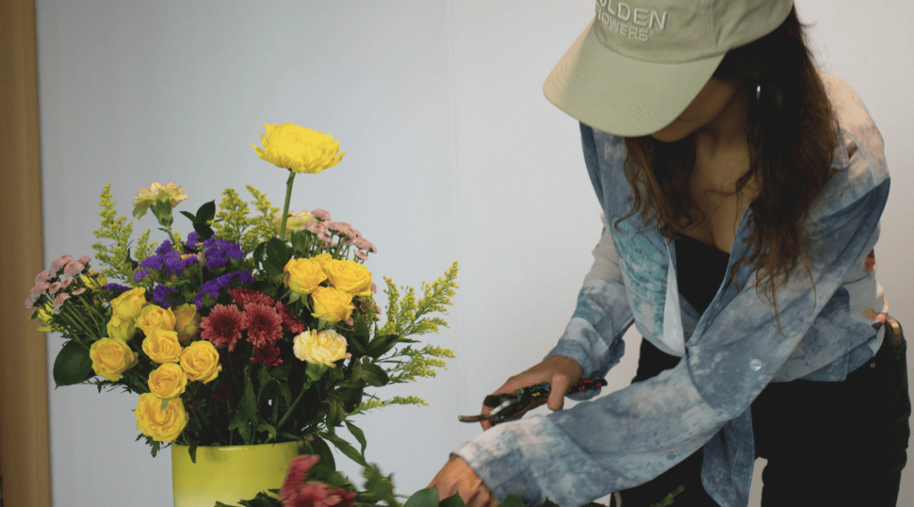 Florist in beige 'Golden Flowers' cap arranging a colorful mothers day bouquet with yellow roses, purple statice, and burgundy chrysanthemums in a yellow vase